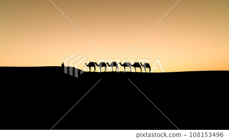 Silhouette of two unidentified Berber men leading a camel caravan across sand dunes during sunset in Sahara Desert, Morocco Silhouette of two unidentified Berber men leading a camel caravan across sand dunes during sunset in Sahara Desert, Morocco 108153496