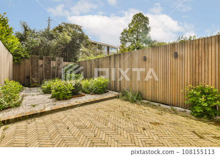 a backyard area with a wooden fence and brick paversed flooring in the fore - image is taken from outside 108155113
