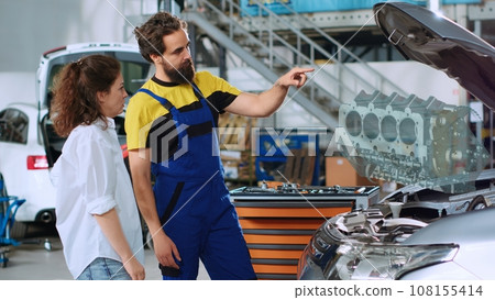 Certified mechanic in auto repair shop using holographic augmented reality to show customer damaged engine inside car. Garage employee using futuristic AR tech to project vehicle parts for client 108155414