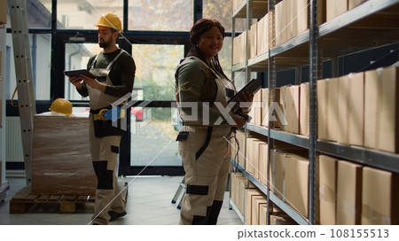 Portrait of joyful warehouse manager holding digital tablet, checking packaging, ensuring smooth operations while employee in background handles packaged goods in cardboard boxes ready to be shipped Portrait of joyful warehouse manager holding digital tablet, checking packaging, ensuring smooth operations while employee in background handles packaged goods in cardboard boxes ready to be shipped 108155513