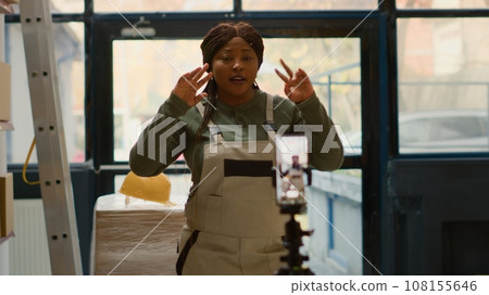 Warehouse employee using cellphone placed on tripod to make training video for interns. Worker filming herself in stockroom showing trainees how to store merchandise, dolly out shot Warehouse employee using cellphone placed on tripod to make training video for interns. Worker filming herself in stockroom showing trainees how to store merchandise, dolly out shot 108155646