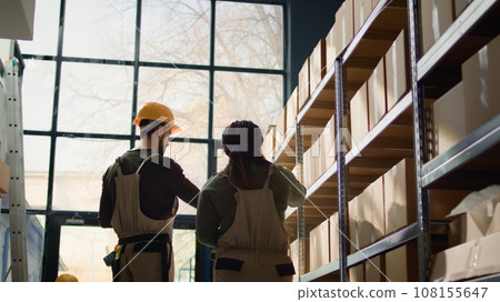 Meticulous assistant in warehouse, scanning items in cardboard boxes on shelves under team leader surveillance, using tablet devices to update prices for online ecommerce website 108155647