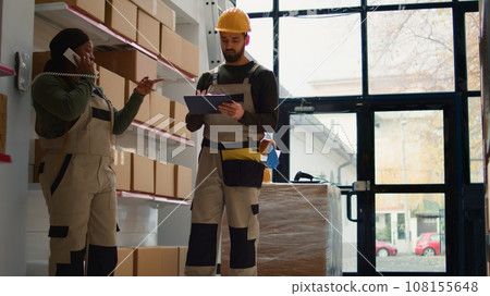 Chief supply chain officer and employee preparing warehouse orders for delivery, receiving phone call in depot while scanning labels on cardboard boxes parcels to be shipped 108155648