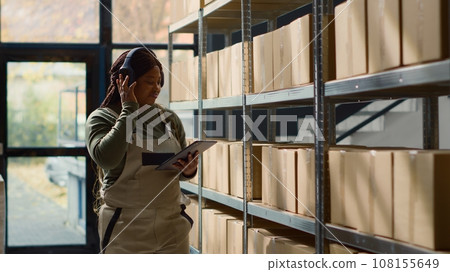 Joyful warehouse chief supply chain officer listening music while inspecting merchandise. Cheerful african american logistics coordinator wearing heaphones while walking through repository rows Joyful warehouse chief supply chain officer listening music while inspecting merchandise. Cheerful african american logistics coordinator wearing heaphones while walking through repository rows 108155649