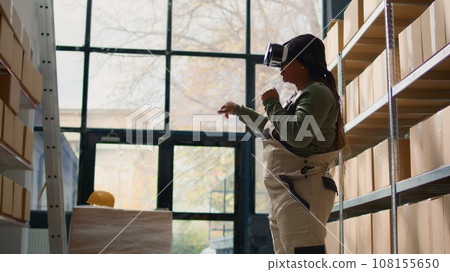 Manager in warehouse wearing VR goggles, using technology to visualize how to best stack cardboard boxes on shelves to make room for more. Supervisor in storage hub uses virtual reality Manager in warehouse wearing VR goggles, using technology to visualize how to best stack cardboard boxes on shelves to make room for more. Supervisor in storage hub uses virtual reality 108155650