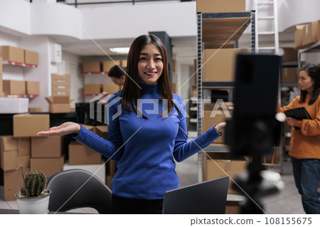 Smiling asian woman recording video in warehouse for retail business development. Entrepreneur filming promotion in storage room explaining customer order picking process Smiling asian woman recording video in warehouse for retail business development. Entrepreneur filming promotion in storage room explaining customer order picking process 108155675