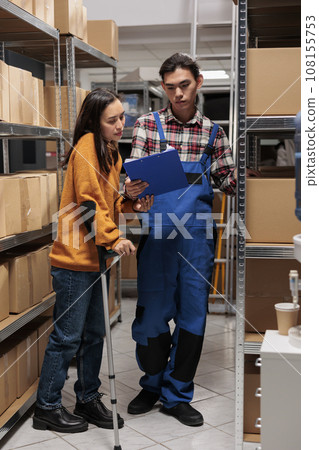 Storehouse asian employees managing inventory optimization in storage room. Warehouse logistics manager on crutches and young man pakcage handler analyzing distribution report on clipboard Storehouse asian employees managing inventory optimization in storage room. Warehouse logistics manager on crutches and young man pakcage handler analyzing distribution report on clipboard 108155753