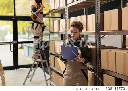 Storage room manager holding clipboard analyzing checklist, looking at clients order before start preparing products for shipping in warehouse. Supervisor wearing protective overall in storehouse 108155770