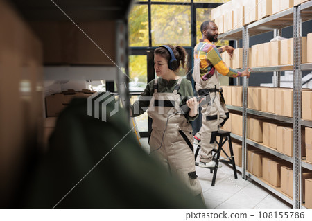 Employee listening music at headphones during inventory in warehouse, scanning cardboard box barcode using store scanner. Storage room supervisor preparing customers orders working at shipping Employee listening music at headphones during inventory in warehouse, scanning cardboard box barcode using store scanner. Storage room supervisor preparing customers orders working at shipping 108155786