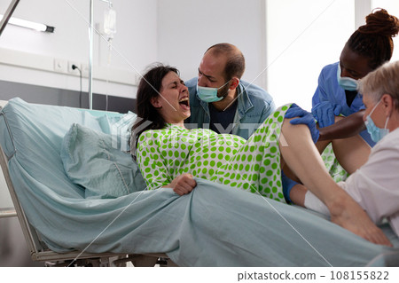 Pregnant woman having caesarean surgery, pushing hard while medical team delivery baby in hopsital ward. Pregnant woman having paniful contractions giving birth to child, being comforting by husband 108155822