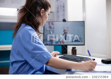 Nurse in videocall taking notes from doctor colleague in professional medical office. Woman writing down information received from general practitioner over telemedicine videoconference Nurse in videocall taking notes from doctor colleague in professional medical office. Woman writing down information received from general practitioner over telemedicine videoconference 108155964
