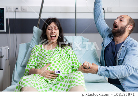 Young couple expecting baby in hospital ward bed at healthcare facility. Woman with pregnancy having painful contractions getting into labor while husband panicking about childbirth. Young couple expecting baby in hospital ward bed at healthcare facility. Woman with pregnancy having painful contractions getting into labor while husband panicking about childbirth. 108155971