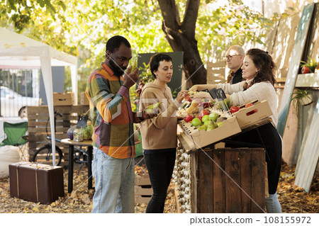 Diverse family tasting fresh organic products and doing shopping at outdoor farmers market stall. Friendly farmer vendor standing behind farm stand offering customers to taste bio apples. Diverse family tasting fresh organic products and doing shopping at outdoor farmers market stall. Friendly farmer vendor standing behind farm stand offering customers to taste bio apples. 108155972