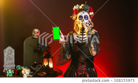 Creepy female model holding greenscreen on smartphone, using mobile phone with isolated mockup template. Woman dressed as santa muerte showing blank chroma key copyspace display. Creepy female model holding greenscreen on smartphone, using mobile phone with isolated mockup template. Woman dressed as santa muerte showing blank chroma key copyspace display. 108155973