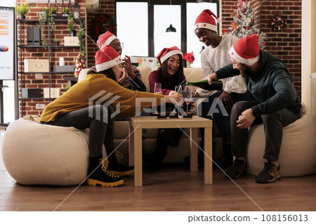 Excited coworkers in santa hats gathered in decorated office and celebrating new year holiday together. Cheerful diverse company employees pouring sparkling wine into glasses 108156013