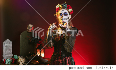 Creepy dead woman trying to lure with hand, reach and tempt victims in studio. Flirty la cavalera catrina model with mexican festival costume on holiday celebration, day of the dead. Creepy dead woman trying to lure with hand, reach and tempt victims in studio. Flirty la cavalera catrina model with mexican festival costume on holiday celebration, day of the dead. 108156017