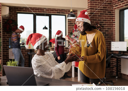 Smiling diverse women colleagues in santa hats exchanging christmas presents at startup company workplace. Cheerful caucasian employee sharing festive gift bag to coworker in decorated office 108156018