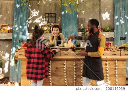 Diverse people looking at farmers market produce on stand, displaying organic fresh fruits and vegetables homegrown. Male vendor showing natural bio products to female client at marketplace. 108156039