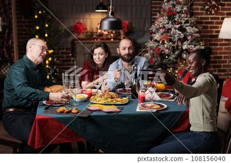 Friends taking pictures on christmas eve, creating beautiful memories of festivity at home. Diverse people having fun with wine and food, enjoying photos for memories at table. 108156040