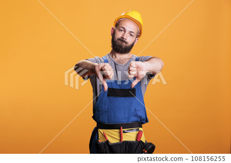 Displeased handyman in overalls showing thumbs down, expressing dislike and disapproval symbol over studio background. Male constructor doing disagreement gesture, being disappointed. Displeased handyman in overalls showing thumbs down, expressing dislike and disapproval symbol over studio background. Male constructor doing disagreement gesture, being disappointed. 108156255