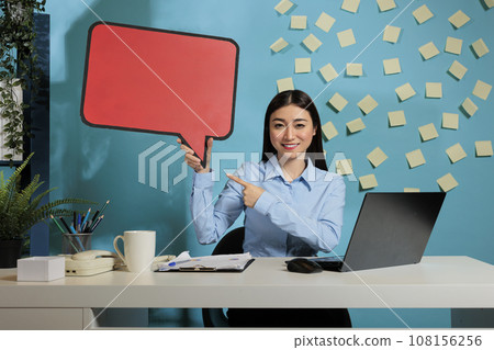 Professional asian woman with advertising speech bubble board against blue wall, showing isolated cardboard text message. Corporate employee holding poster with copy space. Professional asian woman with advertising speech bubble board against blue wall, showing isolated cardboard text message. Corporate employee holding poster with copy space. 108156256