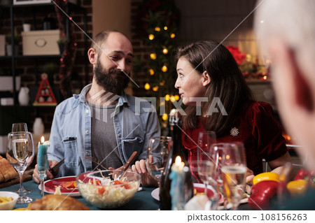 Partners celebrating christmas holiday with friends, acting sweet at dinner table with family. Diverse people enjoying gathering with homemade meal and wine to celebrate xmas eve, winter season. 108156263