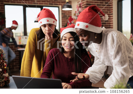 Young women colleagues wearing santa hats discussing and collaborating on tasks on christmas. Diverse smiling coworkers team working on laptop together in decorated office on xmas eve Young women colleagues wearing santa hats discussing and collaborating on tasks on christmas. Diverse smiling coworkers team working on laptop together in decorated office on xmas eve 108156265