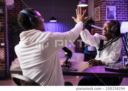 Cheerful online show host high fiving smiling african american guest after successful podcast. Interviewer broadcasting live from living room studio, happy after reaching donations goal Cheerful online show host high fiving smiling african american guest after successful podcast. Interviewer broadcasting live from living room studio, happy after reaching donations goal 108156294