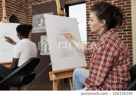 Young woman listening teacher during art workshop learning to draw at beginners group drawing class, creating masterpiece of her own. Stress-relieving creative hobbies for adults Young woman listening teacher during art workshop learning to draw at beginners group drawing class, creating masterpiece of her own. Stress-relieving creative hobbies for adults 108156295
