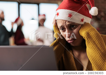 Sad unhappy caucasian woman wearing santa hat working on laptop in office at christmas holiday eve. Exhausted company employee solving task on computer during new year season 108156303