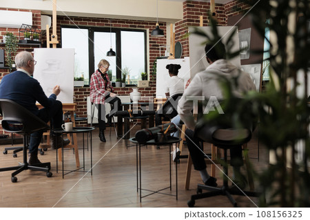 Group of diverse different aged people learning to draw still life objects with proffesional artist, attending group art class, students sitting in classroom drawing with pencil on canvas 108156325