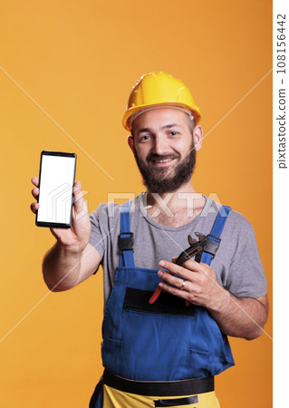 Construction worker holding cell phone with empty screen for advertising in front of camera. Professional builder with hard hat against yellow background in studio shot. 108156442
