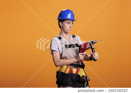 Professional female builder holding drilling gun in new interior renovation project, using power drill nail gun. Woman renovator wearing uniform in studio shot over yellow background. 108156587