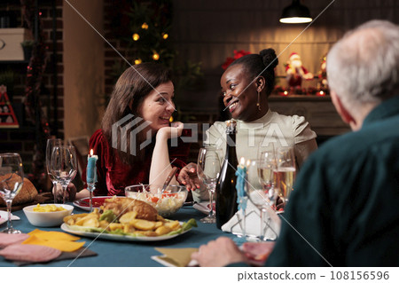 Diverse friends meeting on christmas for dinner, celebrating december holiday at home. People enjoying celebration at table surrounded by festive decorations and xmas tree, feeling happy. Diverse friends meeting on christmas for dinner, celebrating december holiday at home. People enjoying celebration at table surrounded by festive decorations and xmas tree, feeling happy. 108156596
