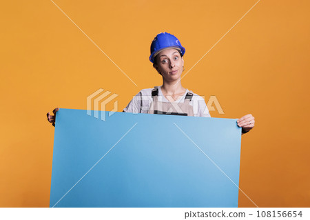 Industrial builder holding blank cardboard sign for advertisement in studio. Optimistic renovating contractor in uniform showing empty carton banner with isolated copyspace, mockup. 108156654
