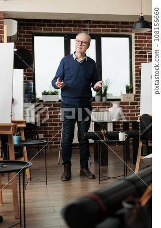 Senior man wearing glasses teaching art to adults in group class, standing in classroom introducing to drawing during creative workshop. Learning to draw from proffesional instructor 108156660