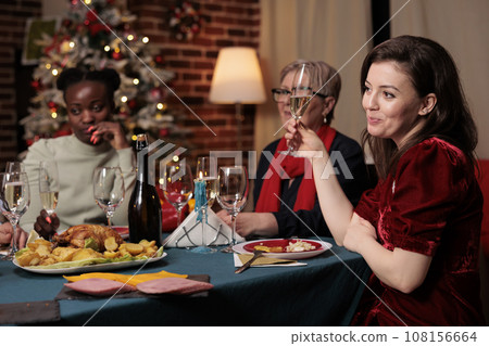 Persons raising glasses for cheers during christmas eve dinner, enjoying family time at home with homemade meal. Diverse people making toast to thank friends for celebrating around the table. Persons raising glasses for cheers during christmas eve dinner, enjoying family time at home with homemade meal. Diverse people making toast to thank friends for celebrating around the table. 108156664