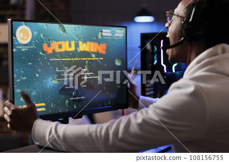 Ecstatic gamer playing intense classic arcade spaceship racing videogame, shaking computer display and yelling after winning. African american man having burst of happiness after game victory 108156755