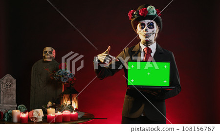 Mexican lady of death showing greenscreen template on laptop, smiling on dios de los muertos. Beautiful woman holding pc with isolated mockup display, chroma key and blank copyspace. Mexican lady of death showing greenscreen template on laptop, smiling on dios de los muertos. Beautiful woman holding pc with isolated mockup display, chroma key and blank copyspace. 108156767