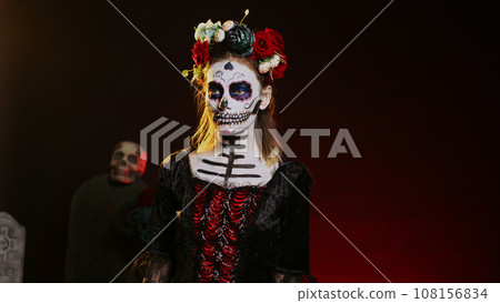 Beautiful lady of dead posing in halloween costume with skull make up to celebrate dios de los muertos on mexican holiday. Spooky woman as santa muerte with black and white body art. Handheld shot. Beautiful lady of dead posing in halloween costume with skull make up to celebrate dios de los muertos on mexican holiday. Spooky woman as santa muerte with black and white body art. Handheld shot. 108156834