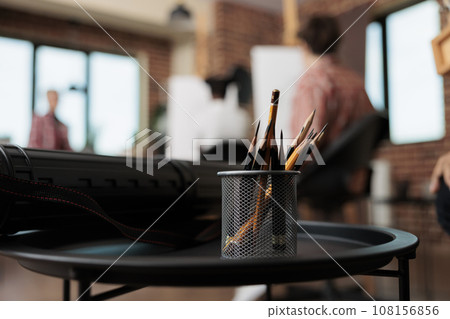 Sketching course. Close up of metal stand holder with graphite pencils standing on table against group of people students sitting at easels on blurred background, studying drawing in art studio 108156856