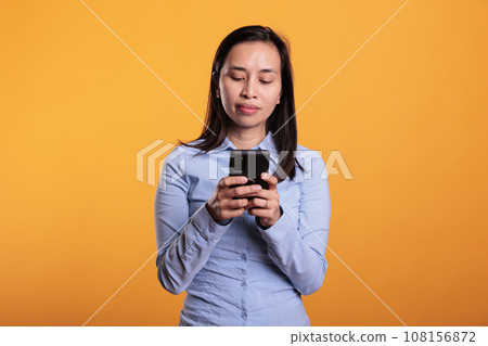 Filipino woman holding mobile phone typing messages, chatting with remote friend in studio over yellow background. Cheerful smiling young adult enjoying sms discussion during break time 108156872