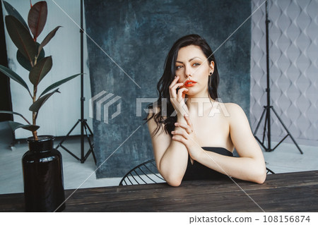 Portrait of young beautiful brunette woman sitting at table on dark gray copy space background. Gorgeous natural beauty girl in cute dress indoors Portrait of young beautiful brunette woman sitting at table on dark gray copy space background. Gorgeous natural beauty girl in cute dress indoors 108156874