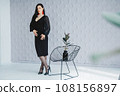 Fashion portrait of a stylish brunette girl wearing a black dress. Woman with long hair wearing red earrings. Model posing standing at the chair. Posing in white studio 108156897
