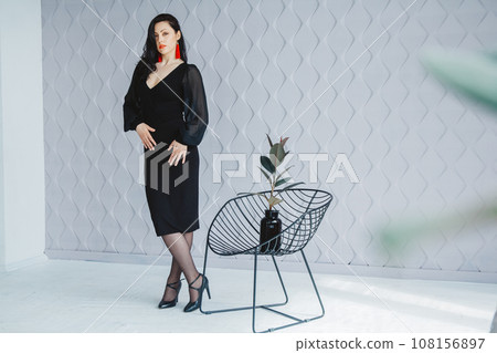 Fashion portrait of a stylish brunette girl wearing a black dress. Woman with long hair wearing red earrings. Model posing standing at the chair. Posing in white studio Fashion portrait of a stylish brunette girl wearing a black dress. Woman with long hair wearing red earrings. Model posing standing at the chair. Posing in white studio 108156897