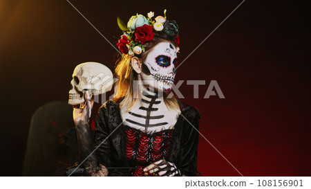 Santa muerte model acting flirty with skull and black roses, wearing traditional body art to celebrate mexican holiday. Looking like goddess of death on holy dios de los muertos ritual. Handheld shot. Santa muerte model acting flirty with skull and black roses, wearing traditional body art to celebrate mexican holiday. Looking like goddess of death on holy dios de los muertos ritual. Handheld shot. 108156901