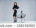 Fashion portrait of a stylish brunette girl wearing a black dress. Woman with long hair wearing red earrings. Model posing standing at the chair. Posing in white studio 108156902