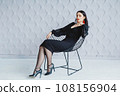 Fashion portrait of a stylish brunette girl wearing a black dress. Woman with long hair wearing red earrings. Model posing standing at the chair. Posing in white studio 108156904