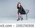 Fashion portrait of a stylish brunette girl wearing a black dress. Woman with long hair wearing red earrings. Model posing standing at the chair. Posing in white studio 108156906