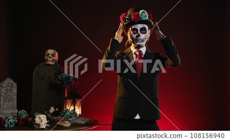 Cavalera catrina with suit and hat looking mysterious wearing festival skull body art, santa muerte celebrating dios de los muertos in studio. Woman looking like holy mexican entity. Cavalera catrina with suit and hat looking mysterious wearing festival skull body art, santa muerte celebrating dios de los muertos in studio. Woman looking like holy mexican entity. 108156940